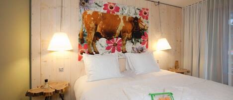 A hotel room with a bed, two bedside lamps, a wooden headboard with a floral print, a small wooden side table, and a green basket with toiletries.