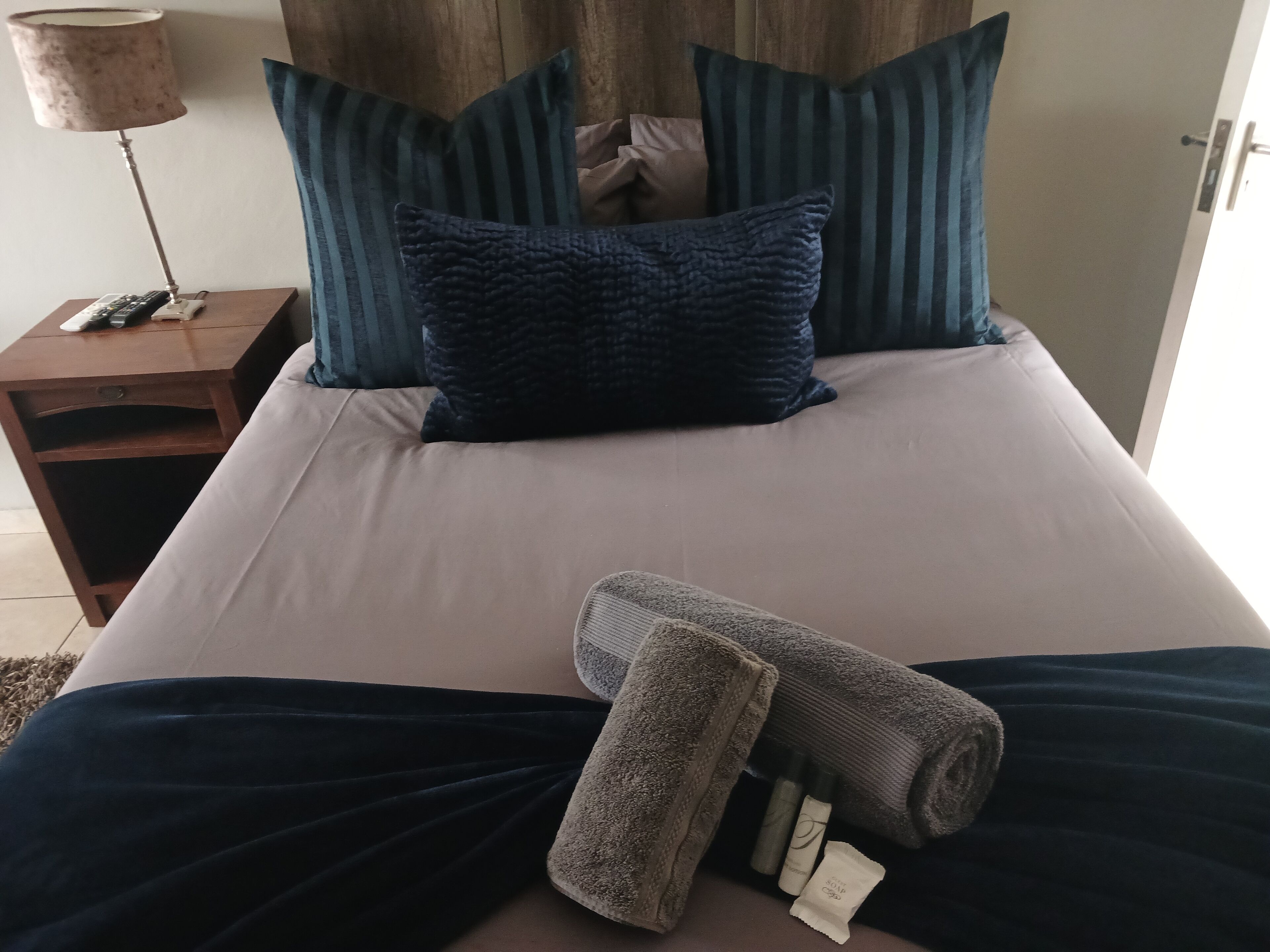 Una cama bien hecha con una colcha azul oscuro, una toalla enrollada y un set de artículos de tocador.