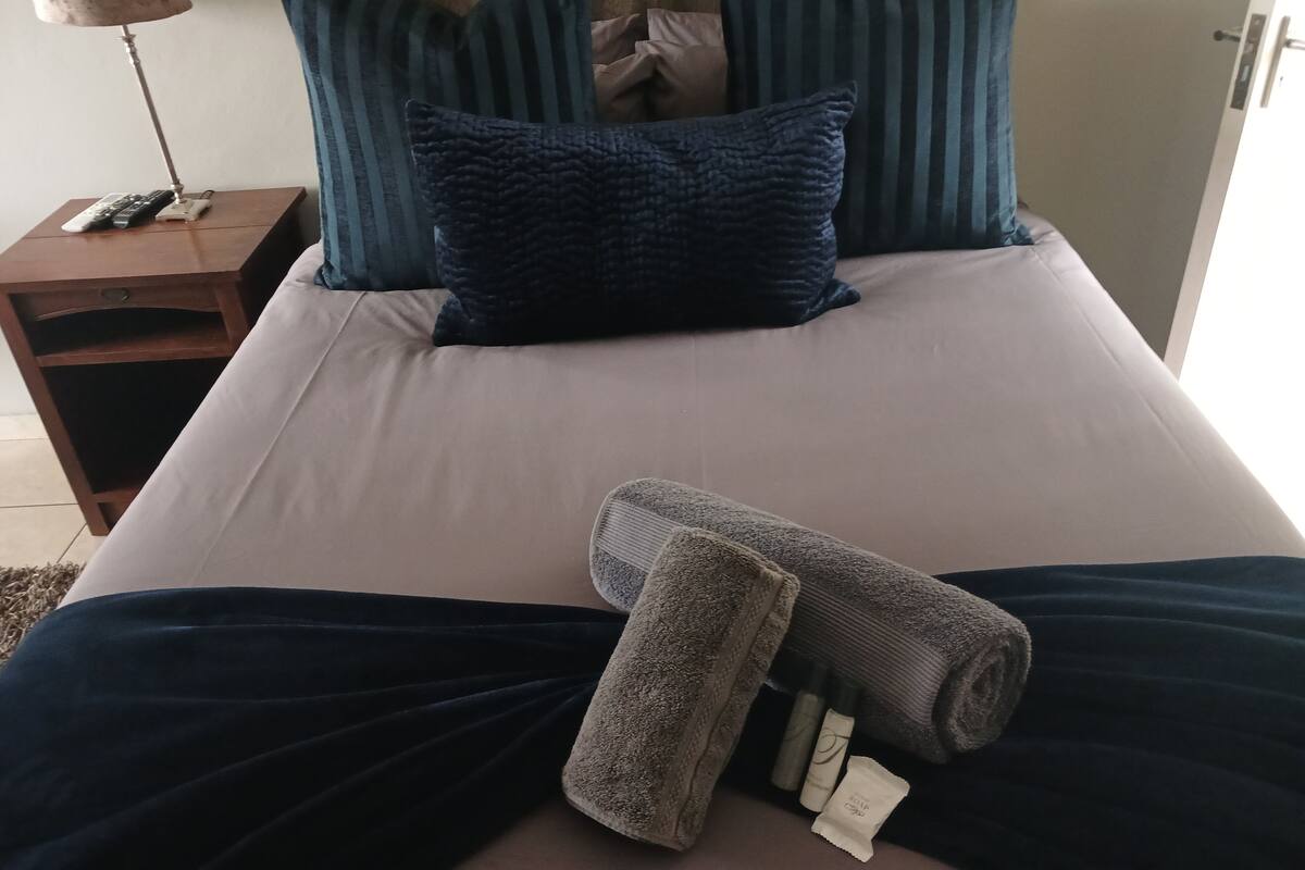 A neatly made bed with a dark blue blanket, a rolled-up towel, and a set of toiletries.