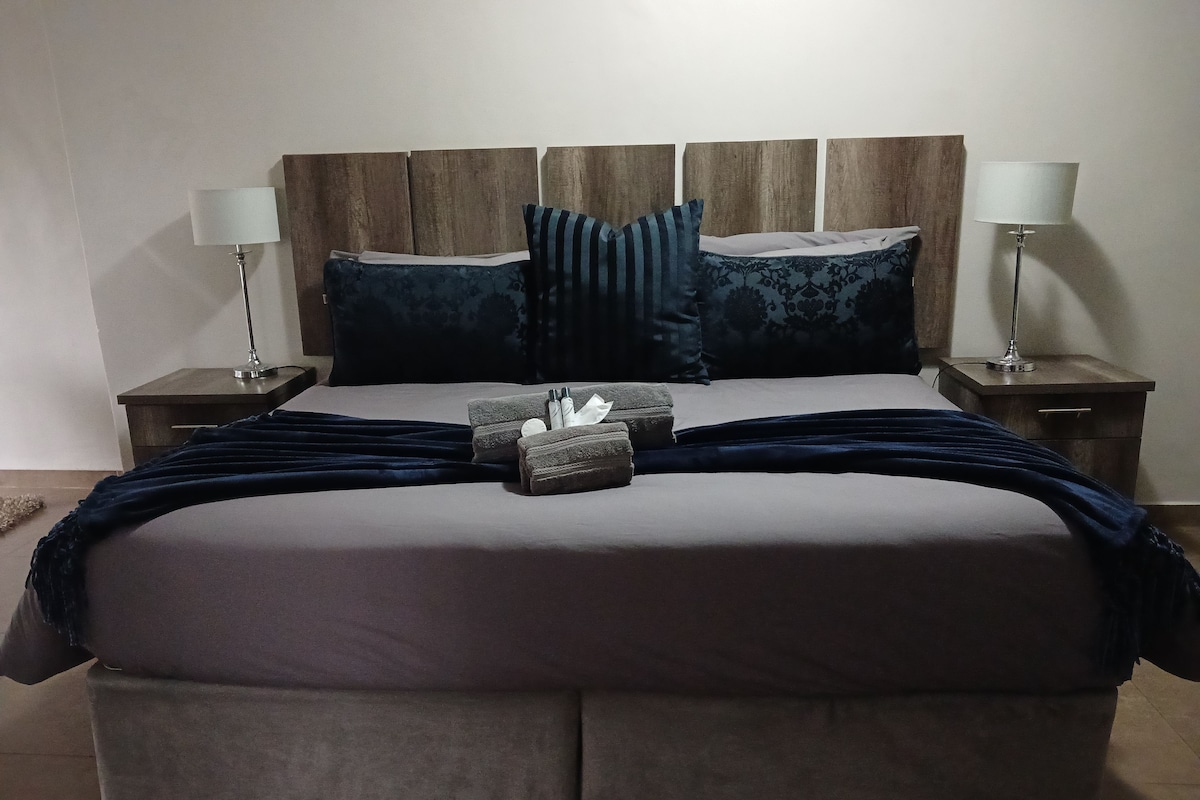 Una cama bien hecha con cabecera, dos mesitas de noche con lámparas y un plaid azul oscuro.