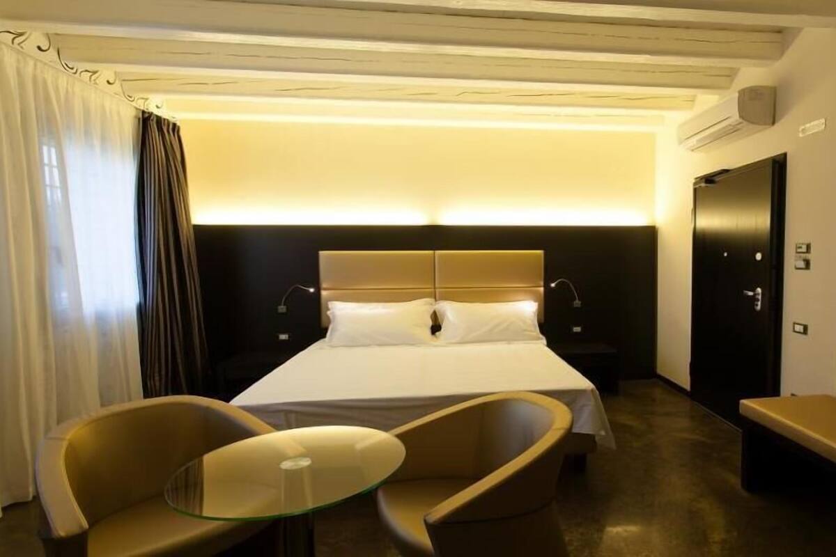 A hotel room with a bed, two chairs, a small table, a bench, and a mirror.