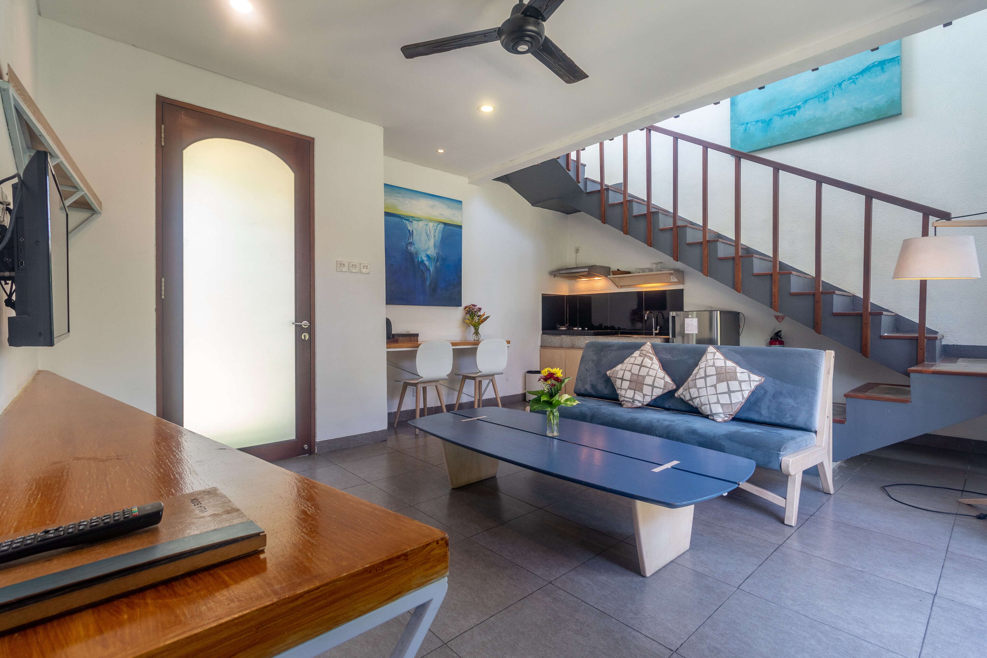 A modern living room with a sofa, coffee table, and a dining area with a wooden table and chairs. A staircase with a railing leads to an upper level.