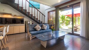 Una sala de estar moderna con sofá, mesa de centro y comedor. Una escalera conduce a una cocina y un balcón con plantas.