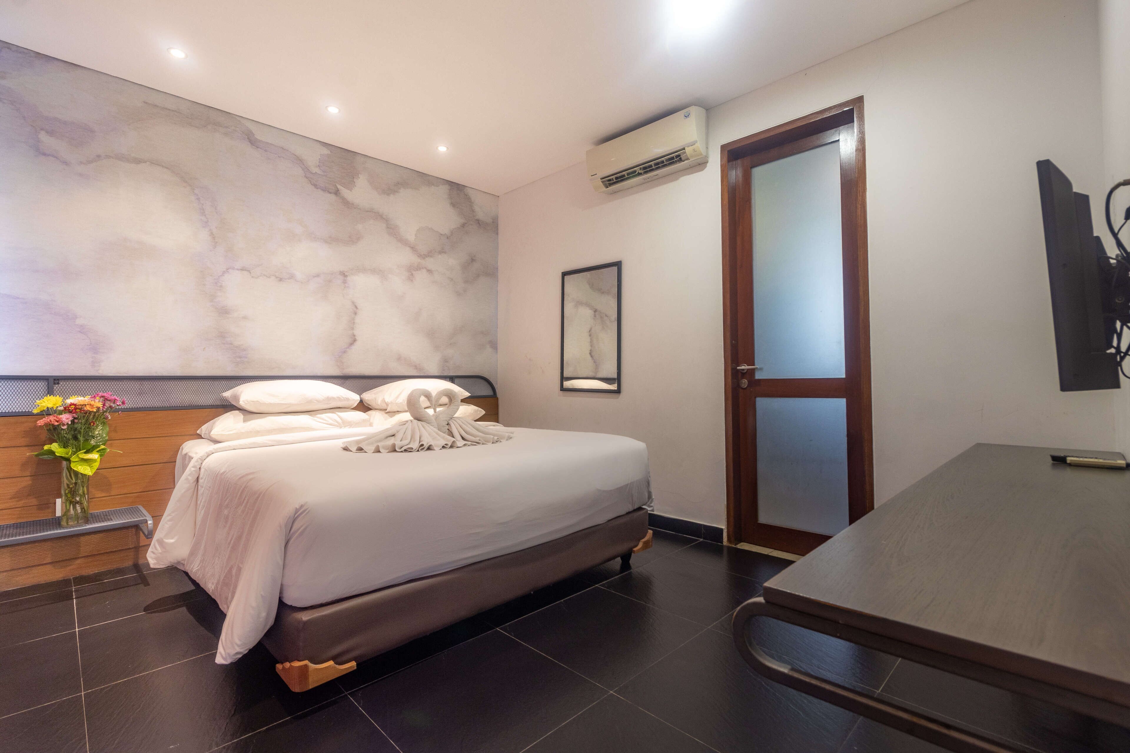 Una moderna camera d'albergo con una parete in marmo, un letto con lenzuola bianche, un comodino in legno con un vaso di fiori e una grande scrivania in legno.