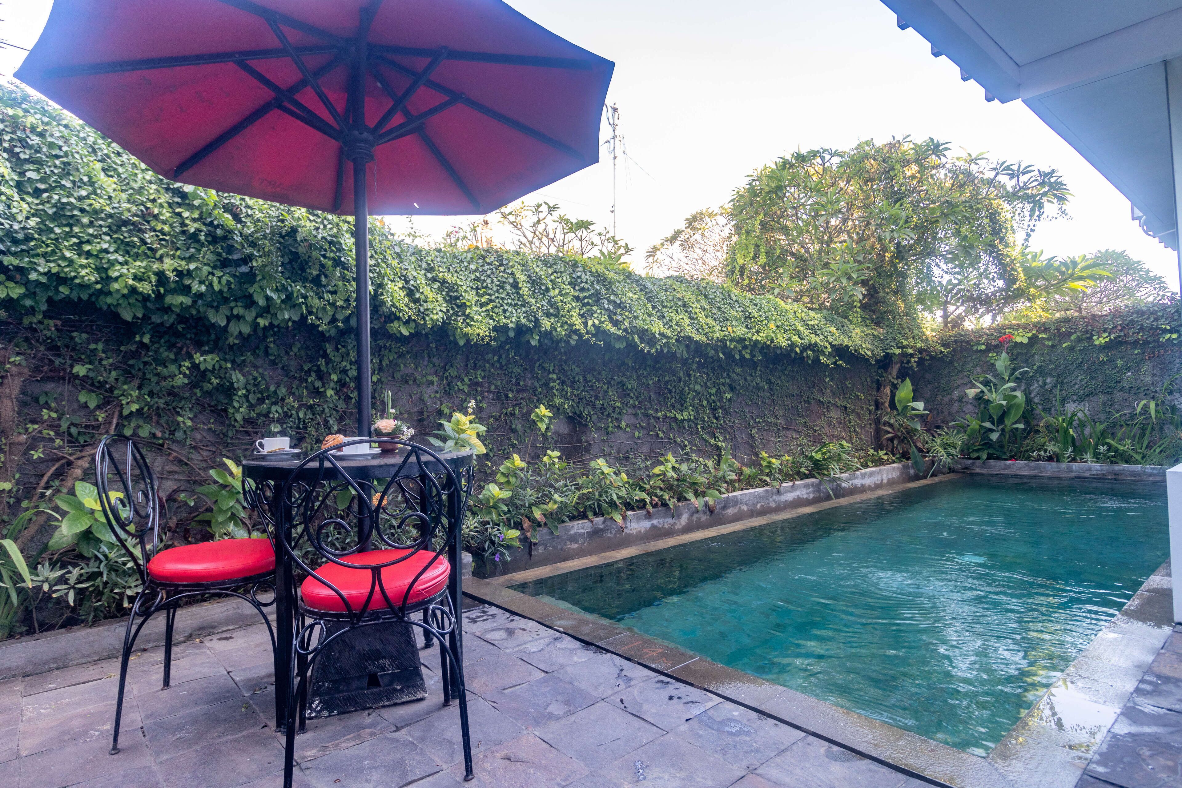 A poolside area with a table and chairs, a red umbrella, and a pool surrounded by greenery.