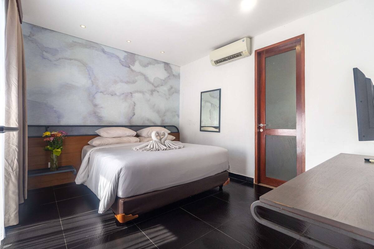 Una habitación de hotel moderna con una cama grande, cabecero de madera, pared de mármol, unidad de aire acondicionado, una puerta y una pequeña mesa.