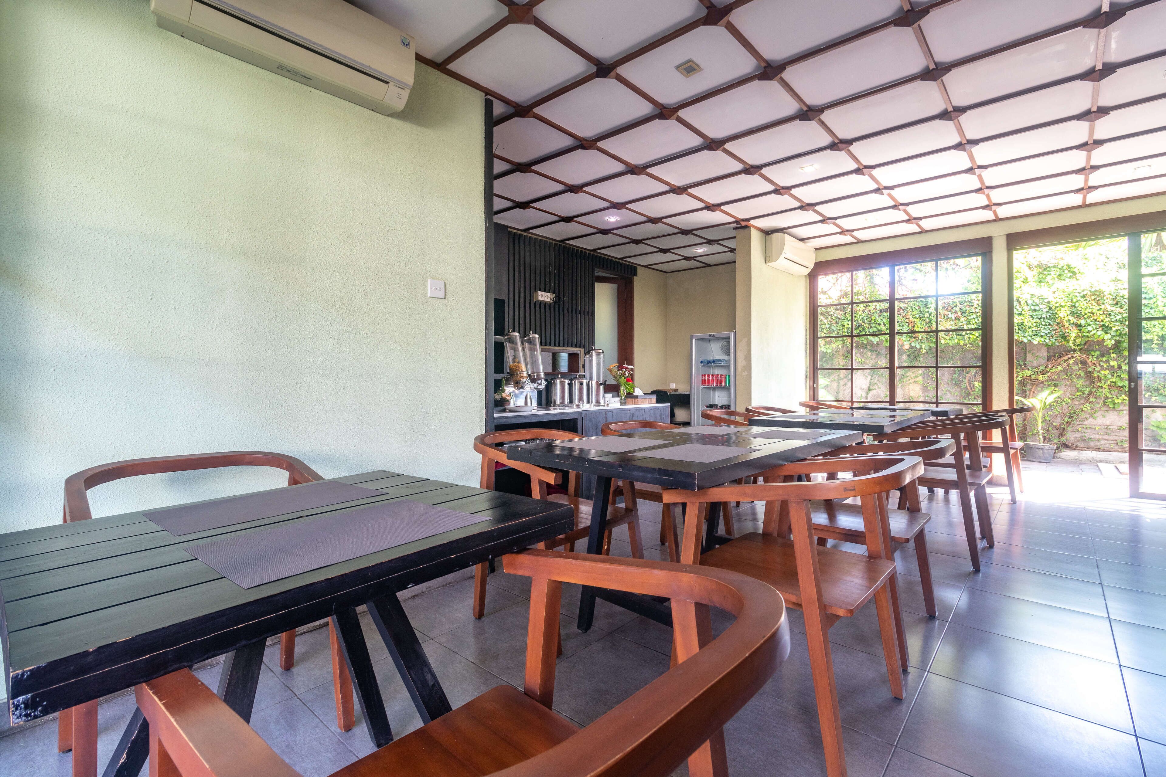 A dining area with multiple tables and chairs, a kitchen area in the background, and large windows allowing natural light.