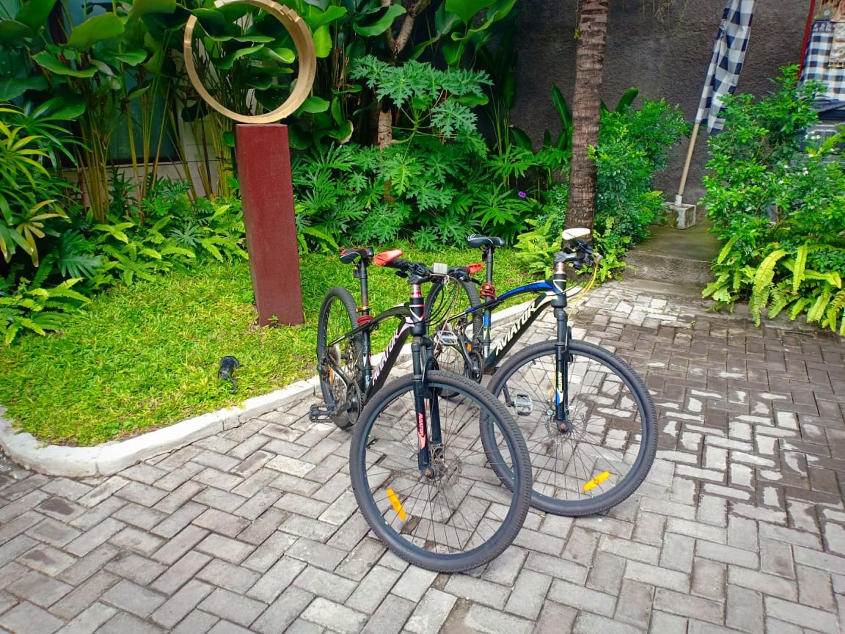 A paved pathway with two bicycles parked on it, surrounded by lush greenery and a wooden structure.