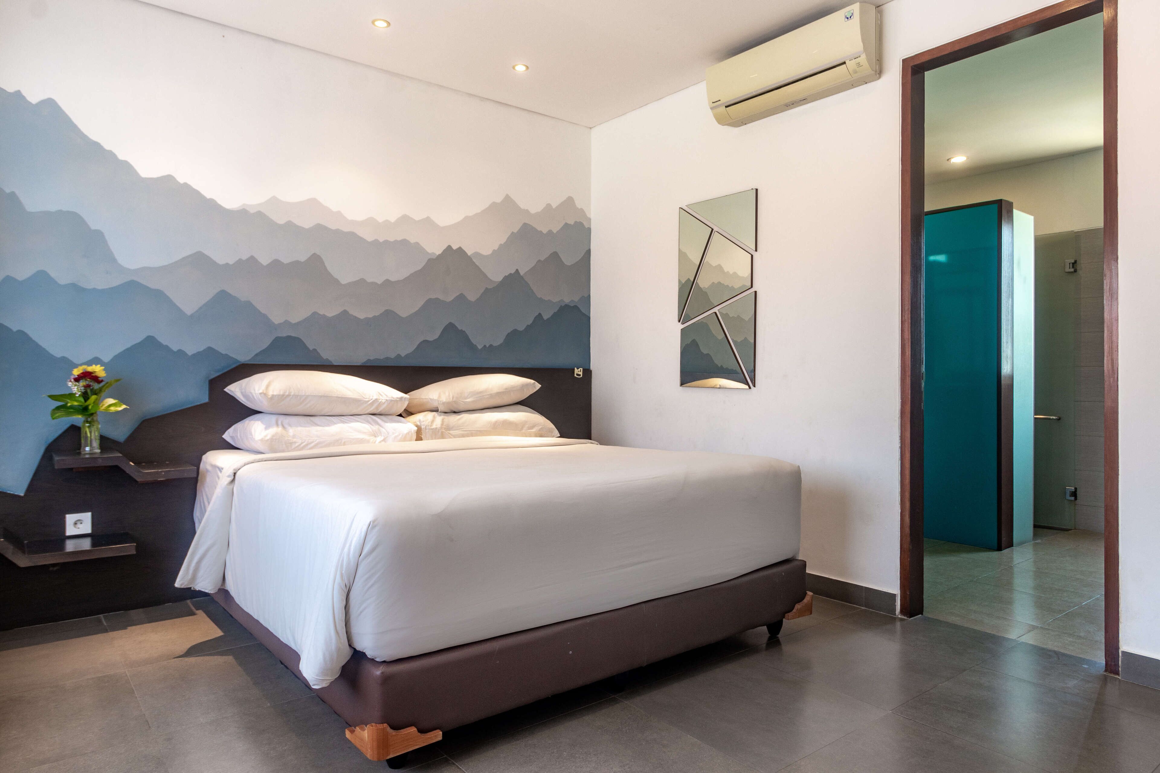 A modern hotel room with a large bed, a mountain-themed wall mural, a small bedside table with a vase of flowers, and a mirror on the wall.