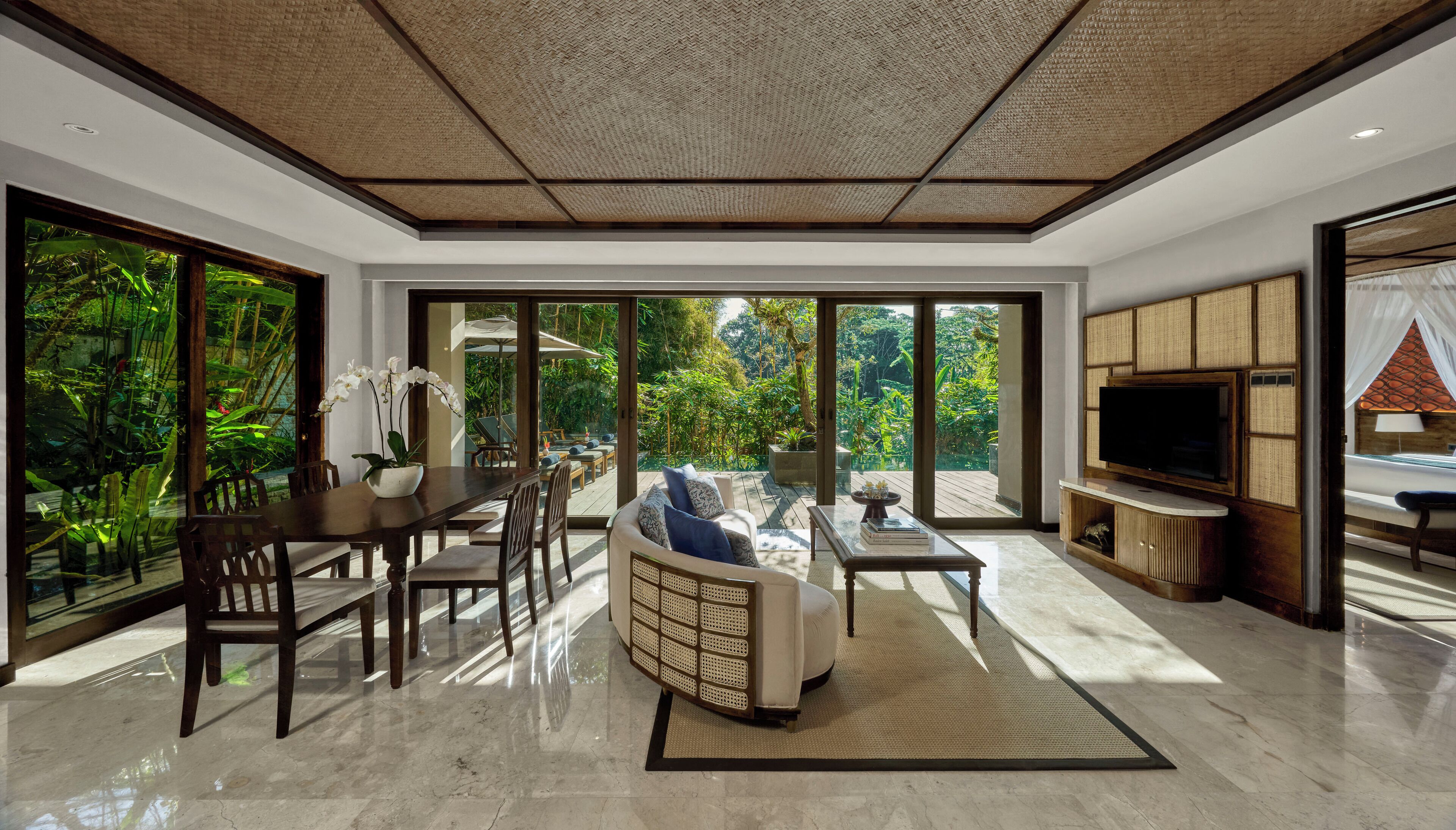 A spacious living area with a dining table, sofa, and TV. Large windows offer views of lush greenery.