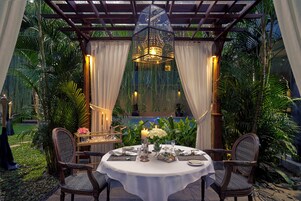 An outdoor dining area with a round table set for two, surrounded by lush greenery and draped curtains.