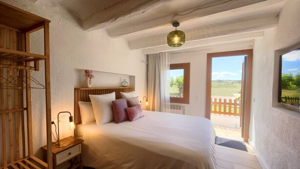 Um quarto com cabeceira de madeira, mesa de cabeceira com luminária, cama com almofadas e vista para o exterior através de uma janela.