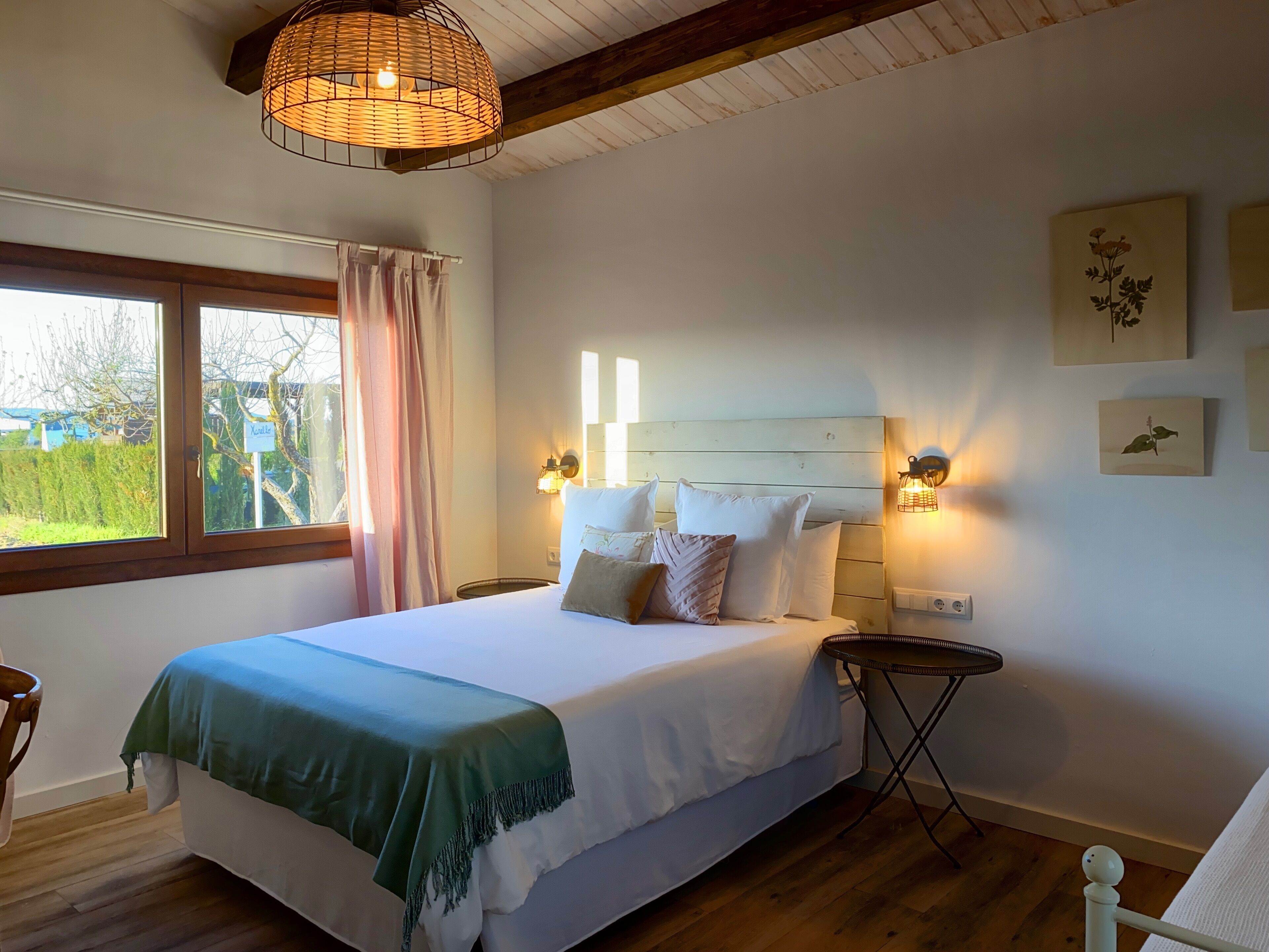 Un dormitorio con cama, mesitas de noche, ventana con cortinas y una obra de arte en la pared.