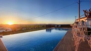 Uma piscina infinita com deck de madeira e grades de metal, com vista para uma paisagem com árvores e colinas ao longe durante o pôr do sol.
