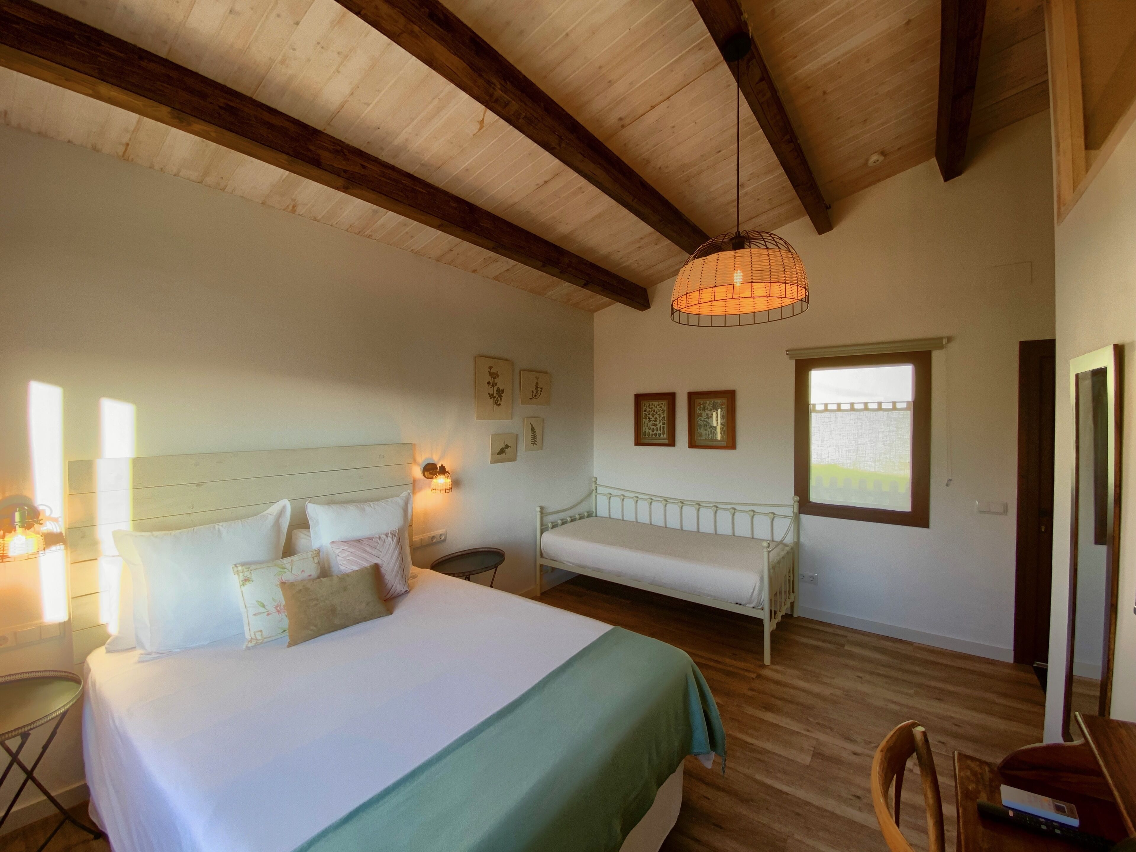 Un dormitorio con una cama grande, un banco, suelo de madera, una lámpara colgante y una ventana.