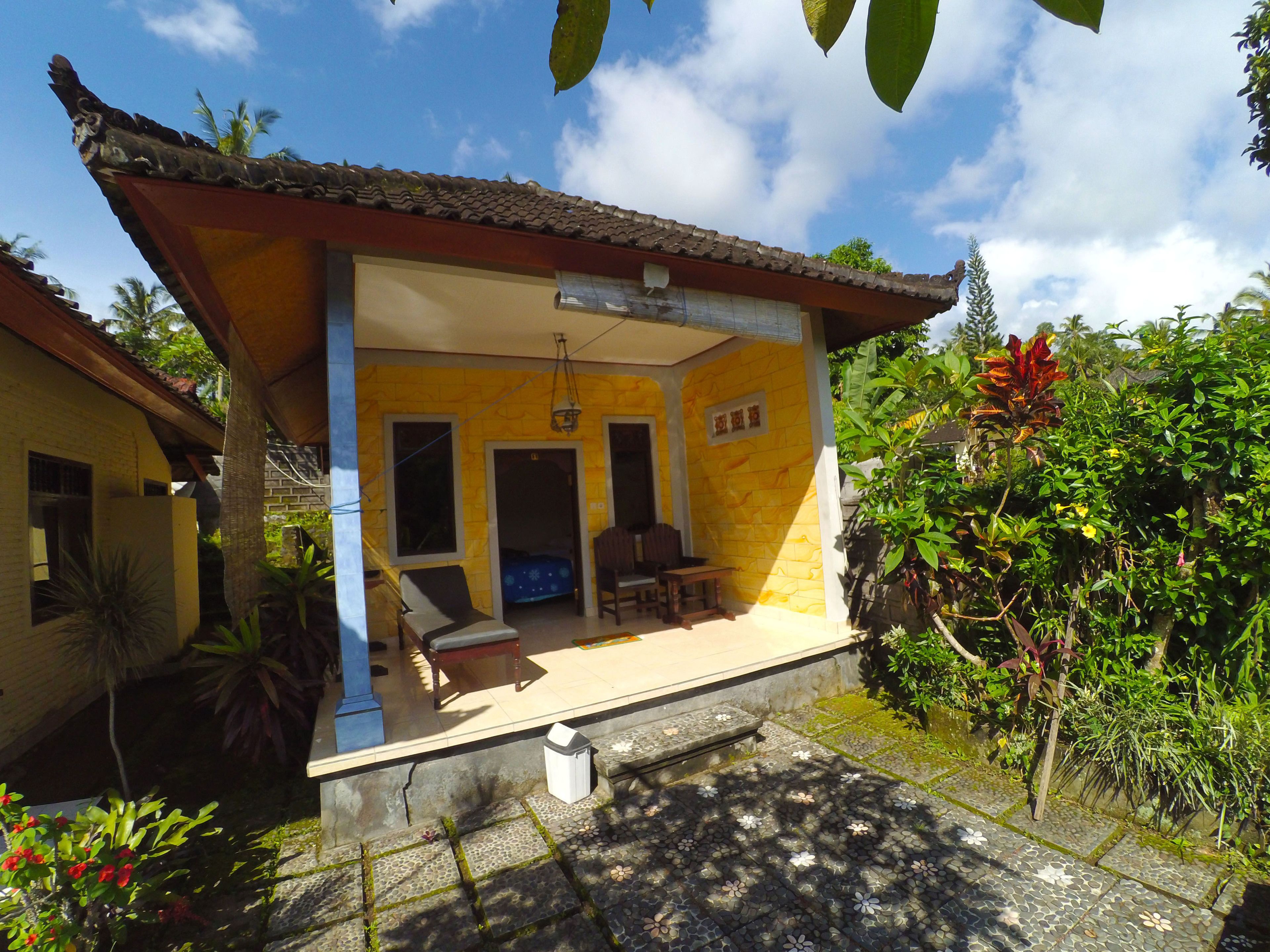 A yellow building with a tiled roof, a covered porch with a bench, and a small garden area with various plants.