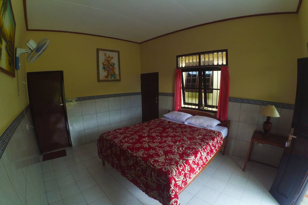 Een slaapkamer met een bed, een raam met gordijnen, een nachtkastje met een lamp en een ventilator aan de muur.