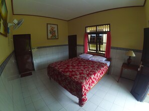 Un dormitorio con cama, ventana con cortinas, mesita de noche con lámpara y ventilador de pared.