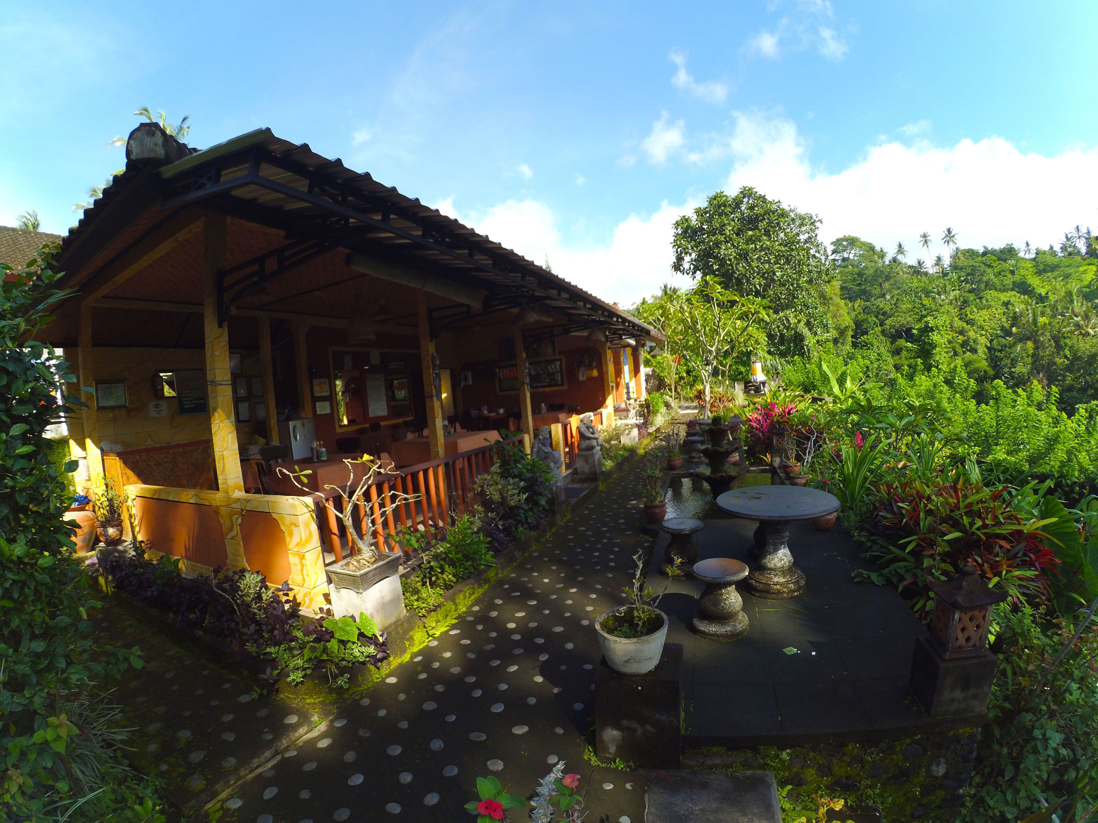 A traditional wooden building with a covered patio, surrounded by lush greenery and a small water feature.