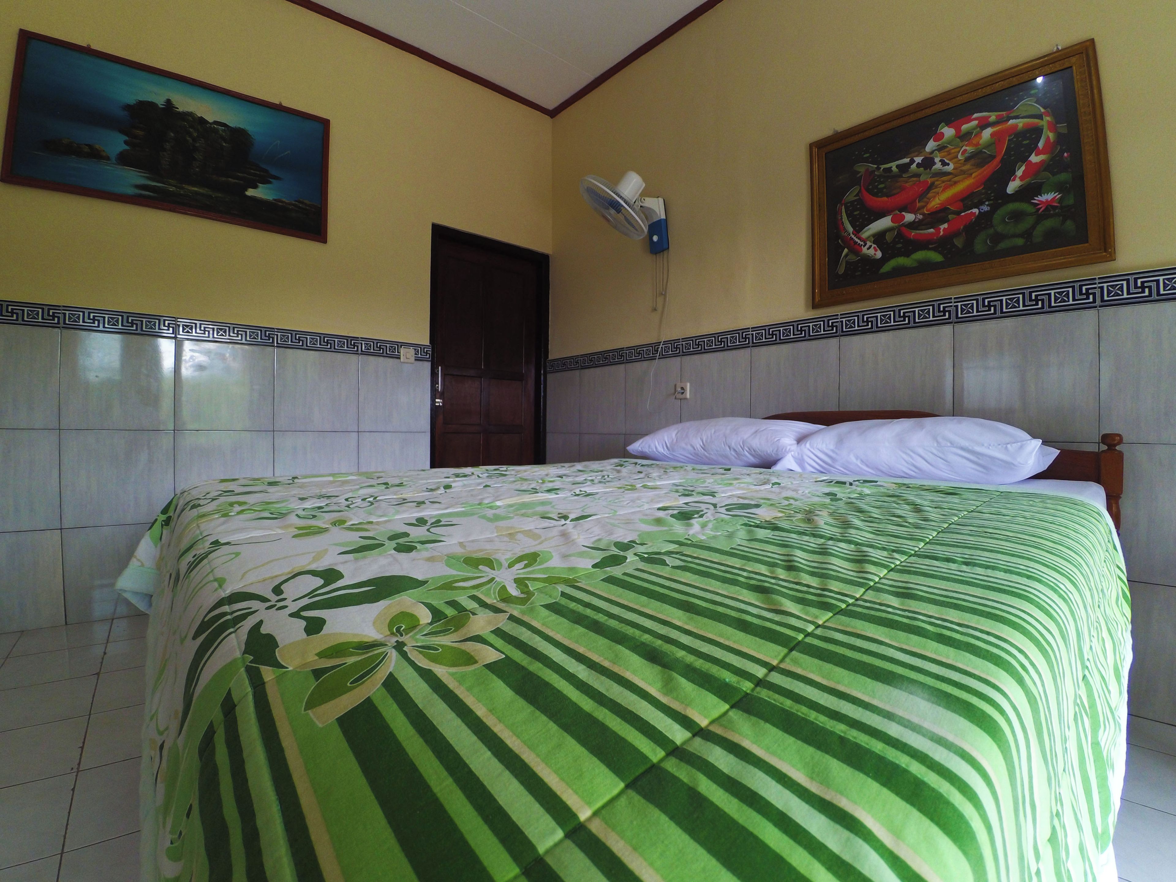 Una habitación con una cama, una puerta, un ventilador de pared y dos cuadros.