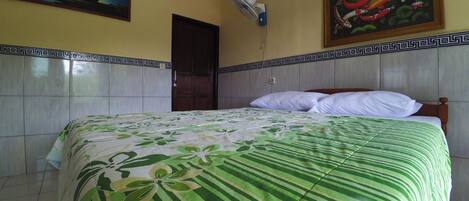 Una habitación con una cama, una puerta, un ventilador de pared y dos cuadros.
