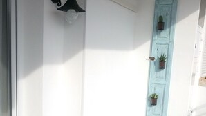Uma pequena varanda com uma mesa preta, duas cadeiras, uma porta azul e um candeeiro fixado na parede.