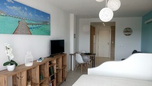 Quarto com cama, TV em estante de madeira, área de refeições com cadeiras e pintura de uma paisagem praiana.