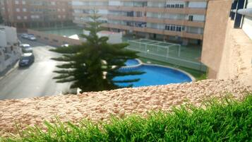Vista de una zona residencial con piscina, una calle con coches y edificios de varios pisos al fondo.