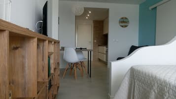 Una habitación moderna con un aparador de madera, una cama blanca, una zona de comedor con mesa y sillas, y una cocina al fondo.