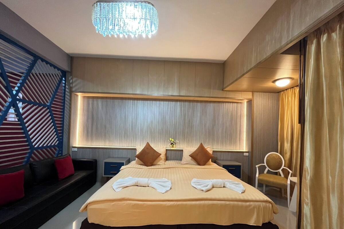 Una habitación de hotel moderna con una cama grande, dos mesitas de noche, una araña y una zona de estar con sofá y sillones.