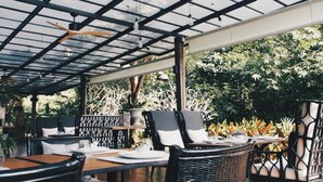 Un área de comedor al aire libre con mesas y sillas, techo de cristal y exuberante vegetación de fondo.