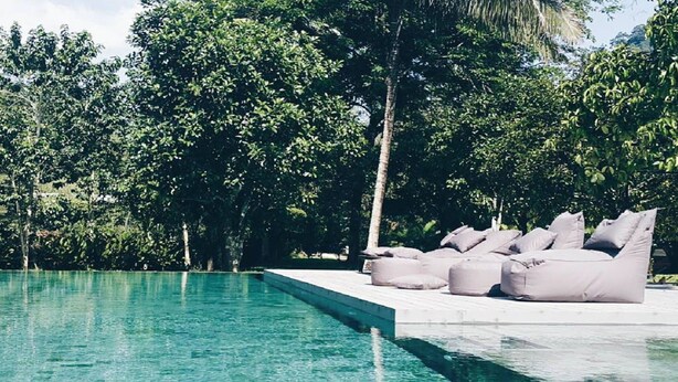Una piscina con agua cristalina, rodeada de abundante vegetación y una terraza con sillones tipo alforzada color beige.