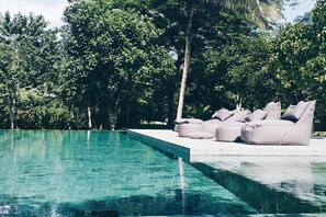 Ein Schwimmbecken mit klarem Wasser, umgeben von üppiger Vegetation und einer Terrasse mit beigefarbenen Sitzsäcken.