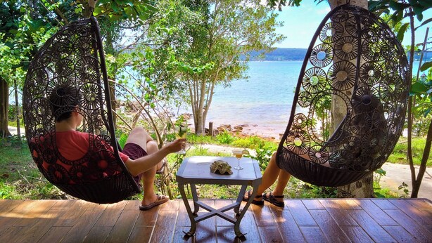 Two people relaxing in wicker swing chairs on a wooden deck with a table, overlooking a beach and water. - Samloem Laguna Resort (Koh Rong Sanloem)