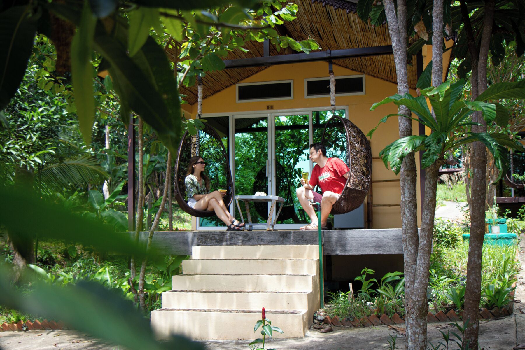 A covered outdoor seating area with a thatched roof, surrounded by lush greenery and a concrete staircase.