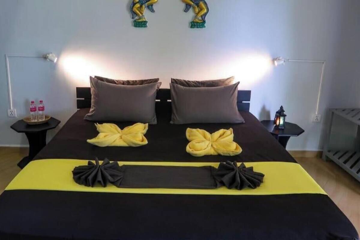 Ein Doppelbett mit einer schwarz-gelben Tagesdecke, zwei braune Kissen und dekorative schwarz-gelbe Schleifen. Zwei Nachttische mit Lampen, einer kleine Statue und zwei Flaschen stehen auf einem Tisch.