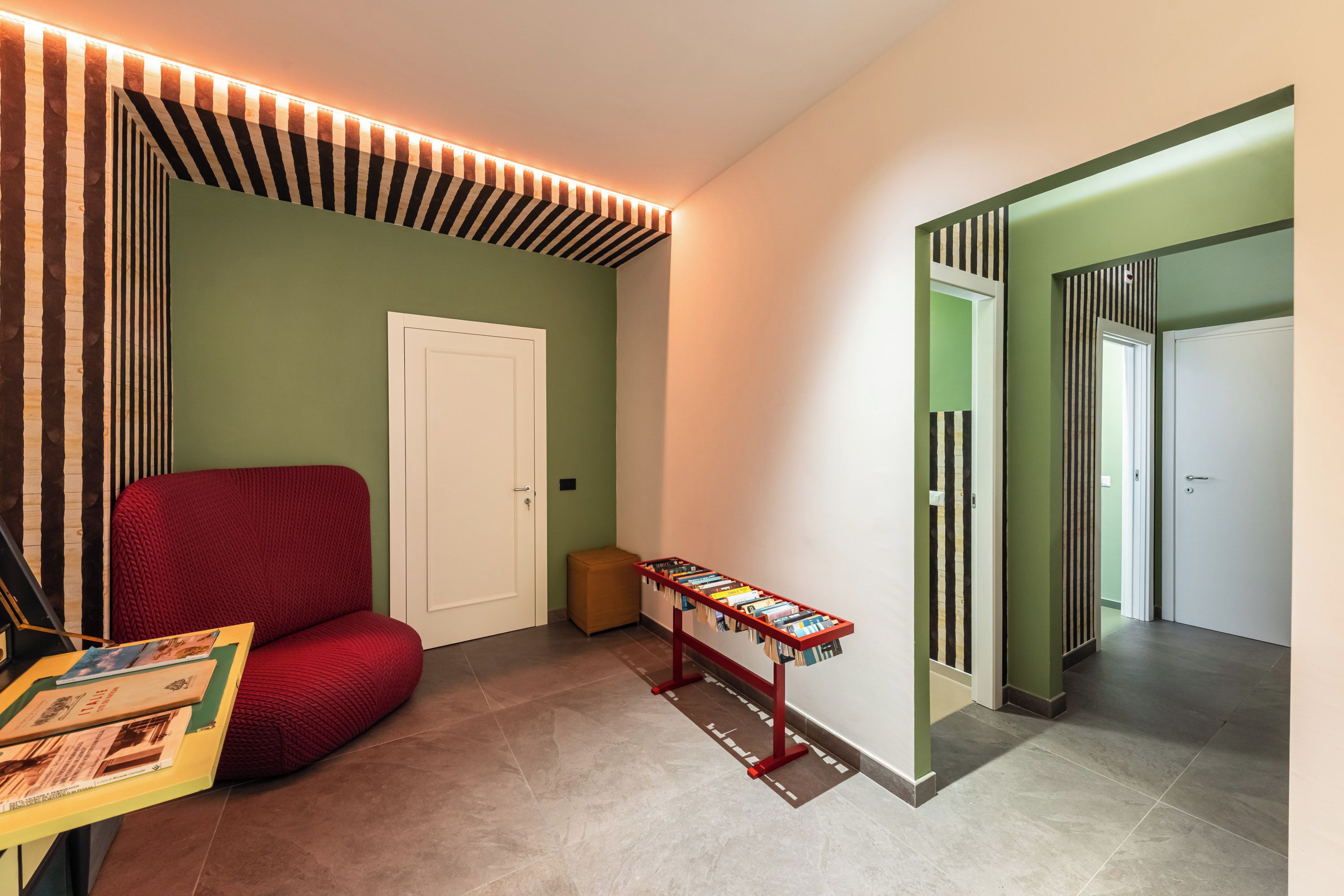 A hallway with a red upholstered chair, a small table with books, and a door with striped wallpaper.