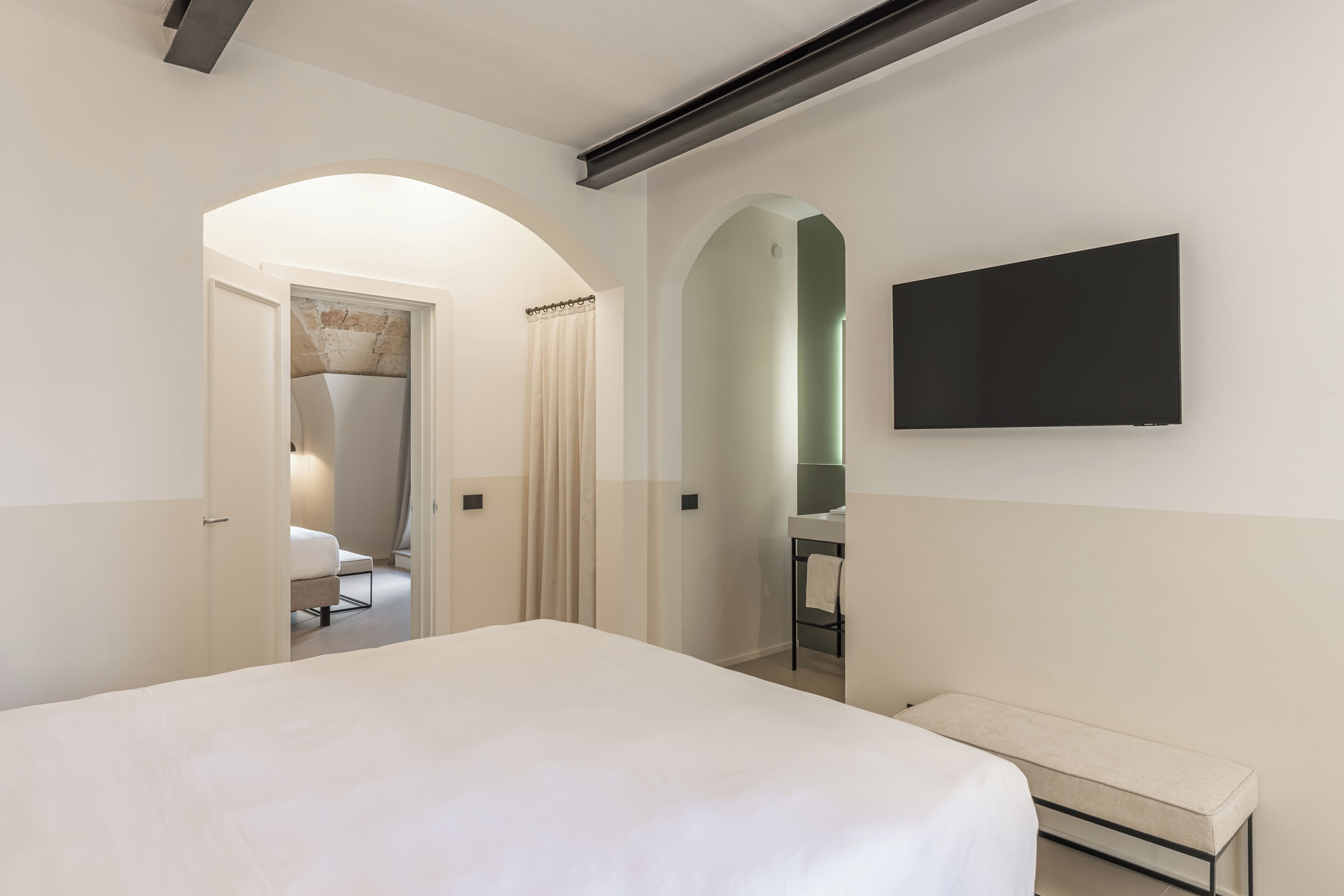 A modern hotel room with a large bed, a bench, a TV mounted on the wall, and a bathroom visible through an archway.