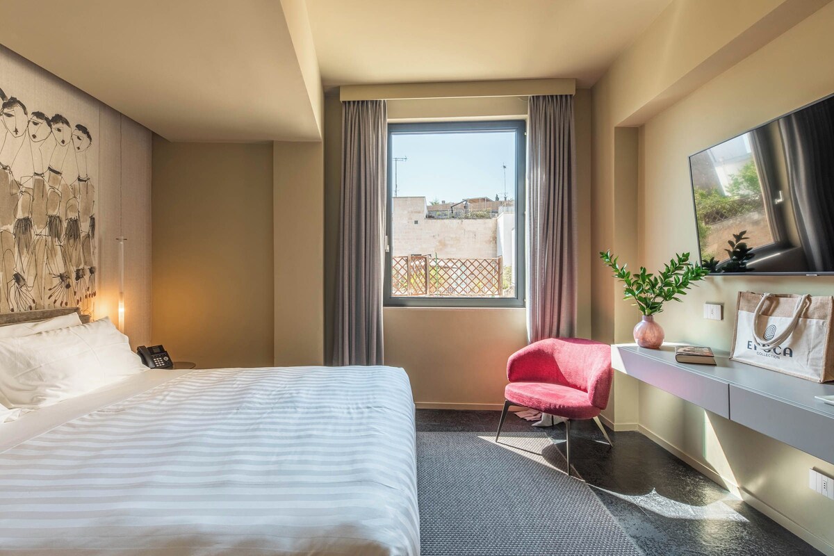 Une chambre d’hôtel comprenant un lit, un bureau avec une chaise, une télévision, une fenêtre avec des rideaux et une plante.