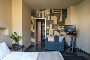 Habitación de hotel con una cama, una mesita con teléfono y una planta, un sofá, un televisor y una bicicleta estática.