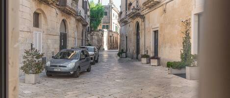 A narrow cobblestone street lined with historic buildings, parked cars, and potted plants.