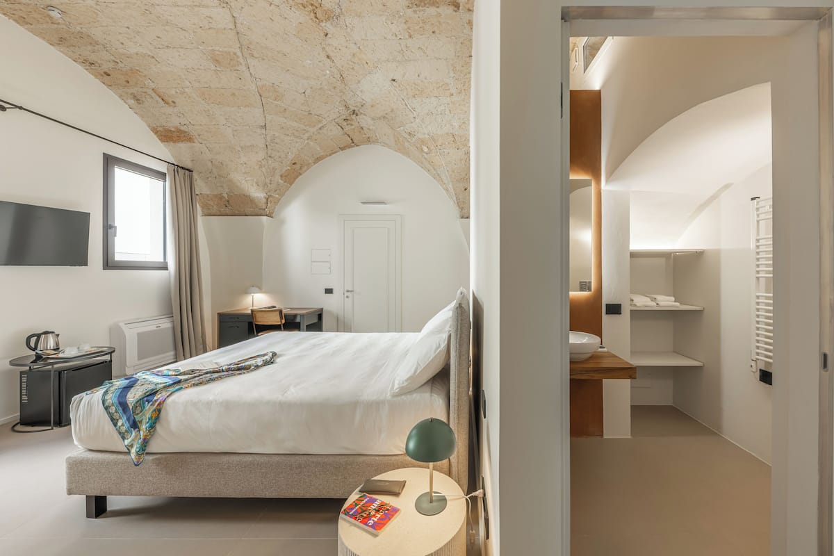 A modern bedroom with a large bed, a bedside table with a lamp, a window, and a bathroom visible through an open door.