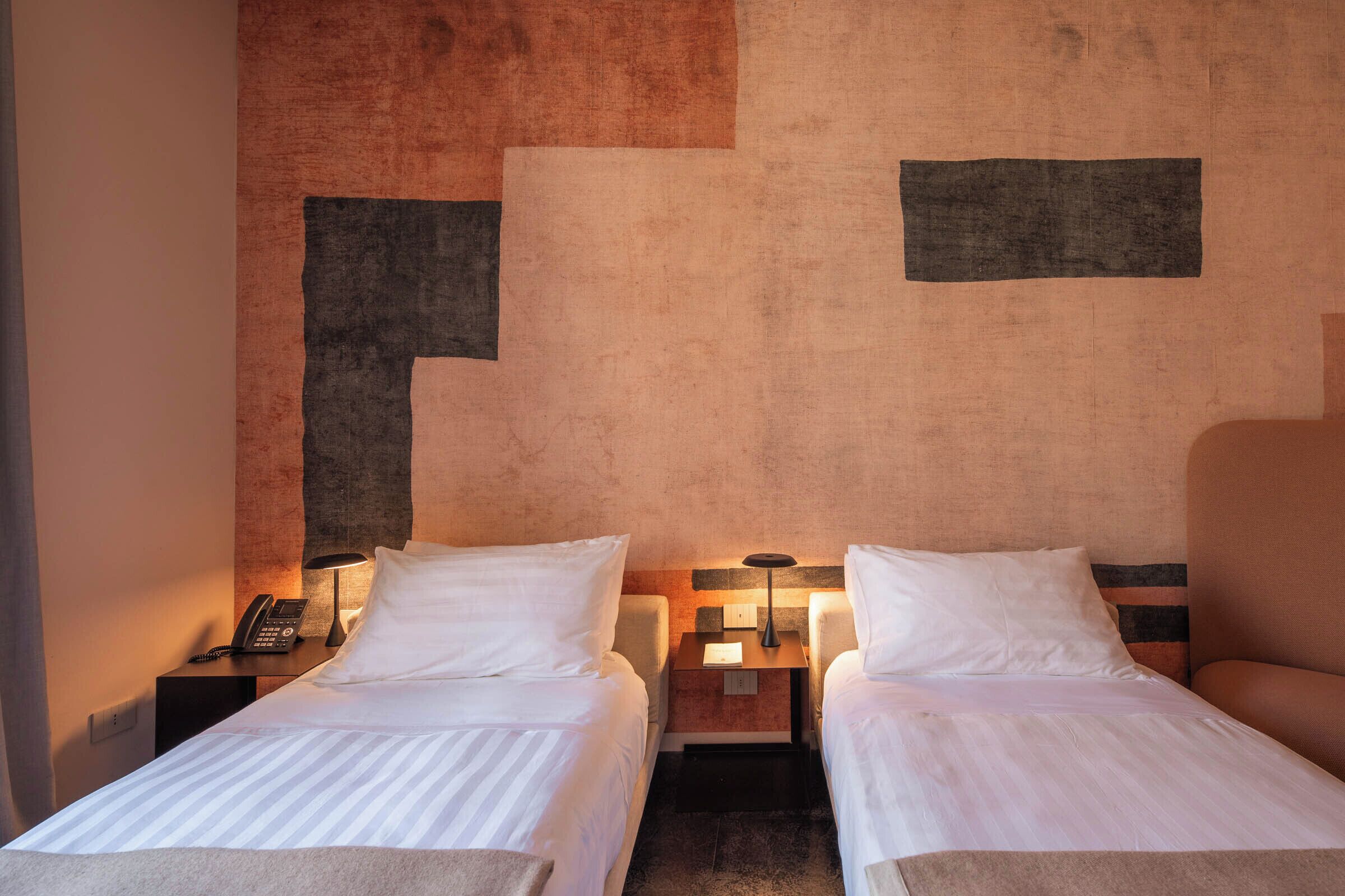 A hotel room with two single beds, bedside tables with lamps, a phone, and a small nightstand.