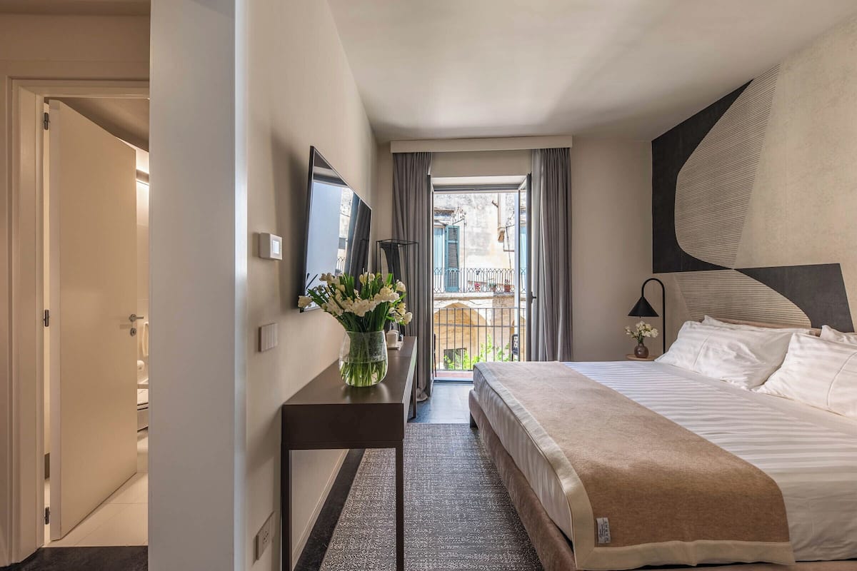 A hotel room with a large bed, a bedside table with a vase of flowers, a flat-screen TV, and a view of the outside balcony.