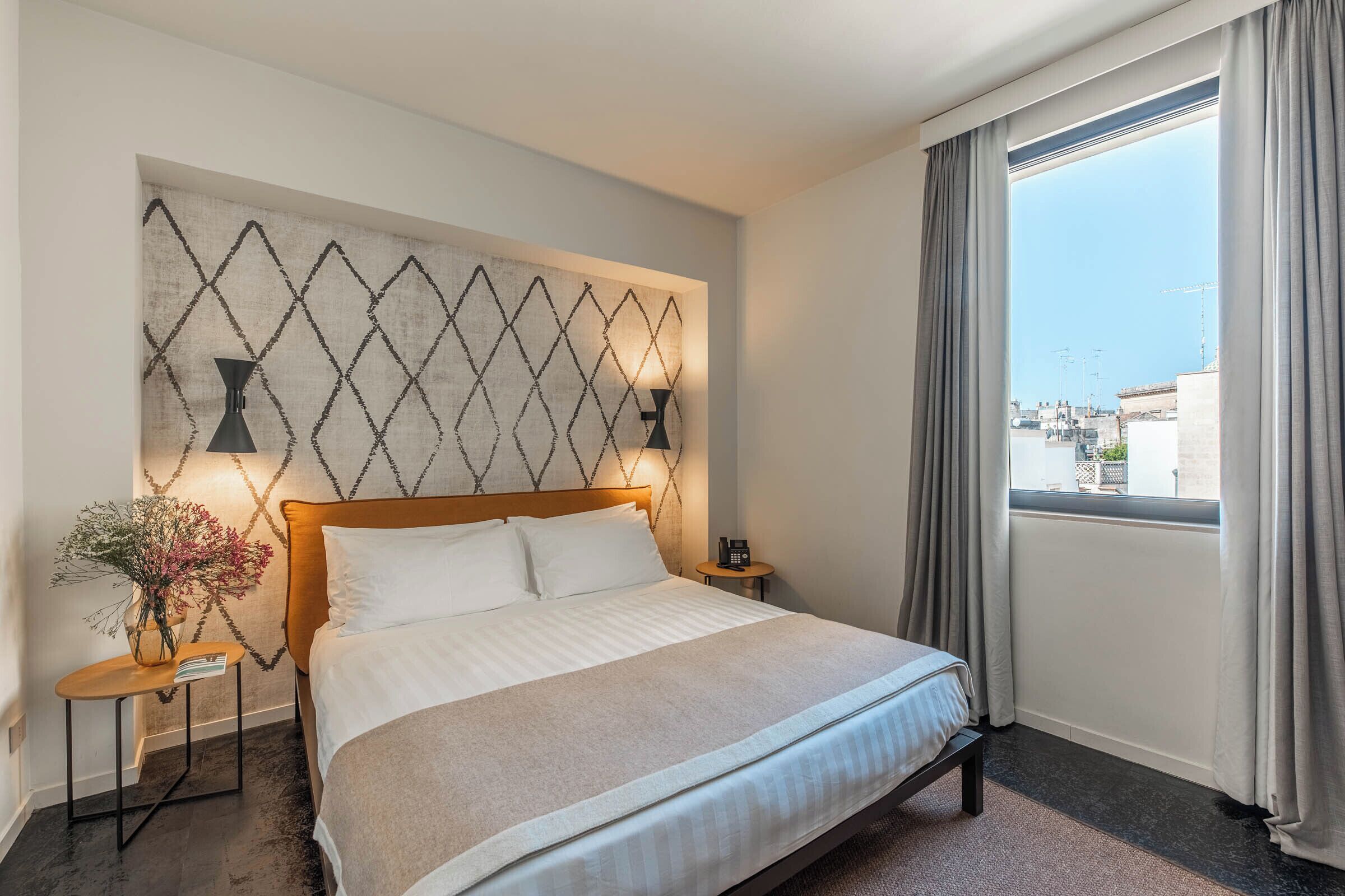 Une chambre d’hôtel avec un grand lit, une table de chevet avec une lampe, un vase de fleurs et une vue sur la ville par la fenêtre.