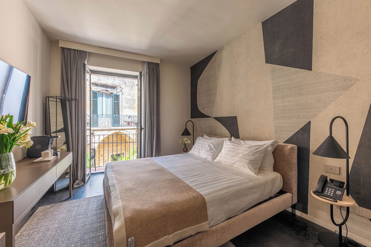 Quarto de hotel com cama, mesa de cabeceira, abajur e vista para a varanda externa.