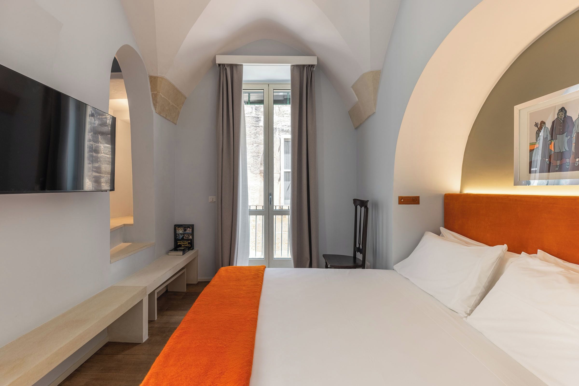 A hotel room with a large bed, a flat-screen TV mounted on the wall, a bench, and a framed picture on the wall.