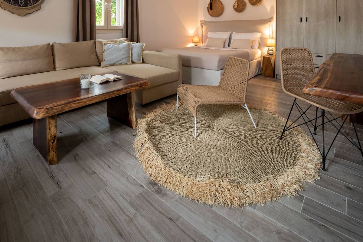 A cozy hotel room with a wooden floor, a sofa, a round woven rug, a bed with white linens, and a wooden wardrobe.