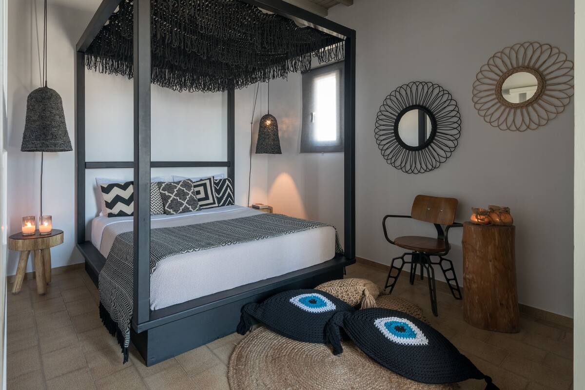 A four-poster bed with a black frame and white bedding, a woven rug, and a round mirror on the wall.