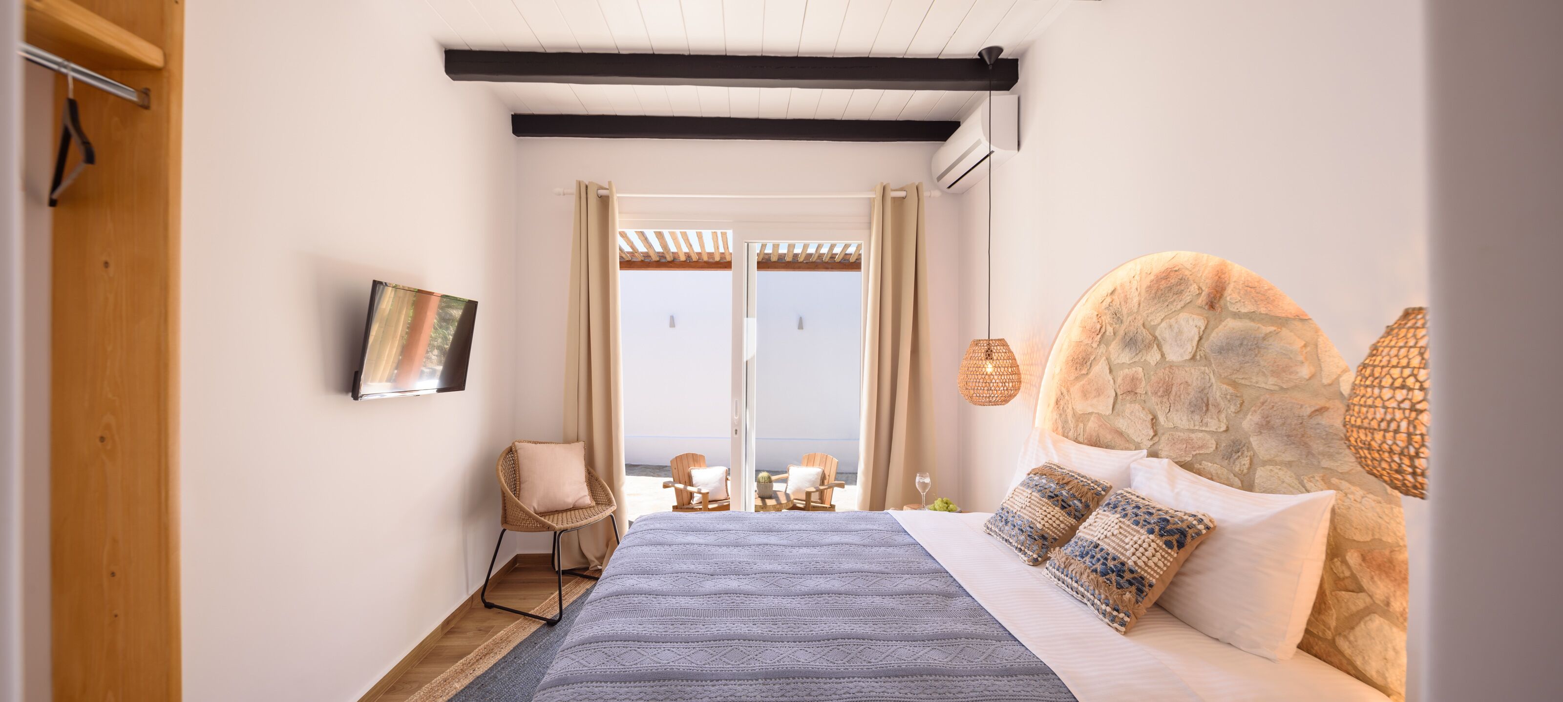 Un dormitorio con cabecera de piedra, una cama con manta azul, una silla de mimbre y vistas al mar a través de un ventanal.